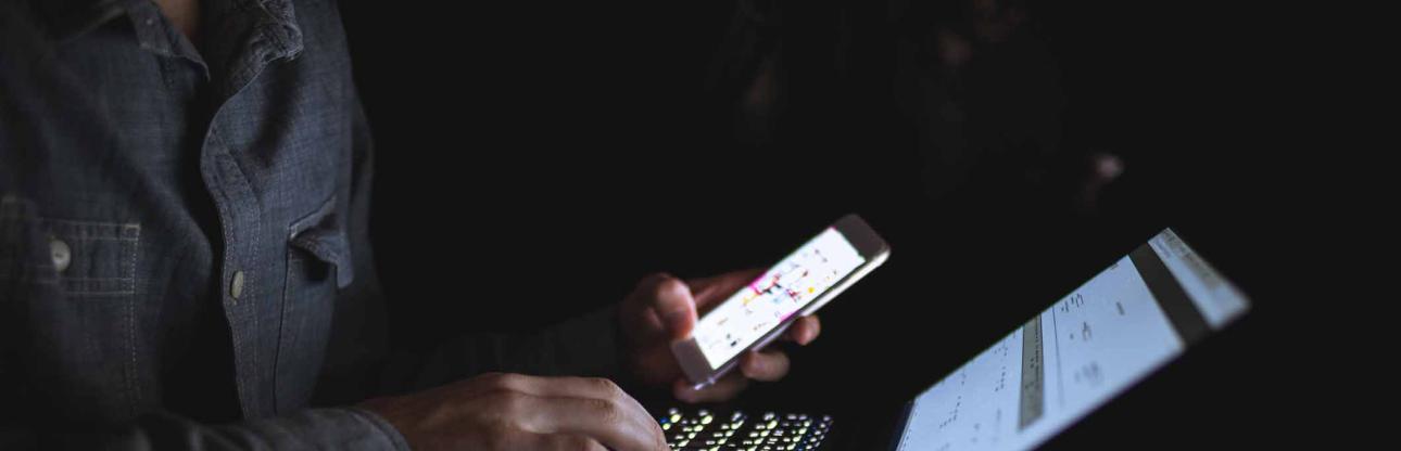 Person using a laptop keyboard in a dark room, while holding and looking at a smartphone.